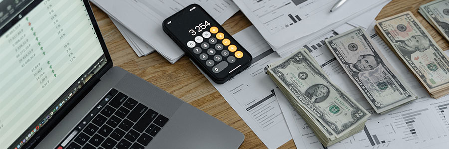 A top view of a desk scattered with an open laptop, calculator, pen, cash and accounting documents.